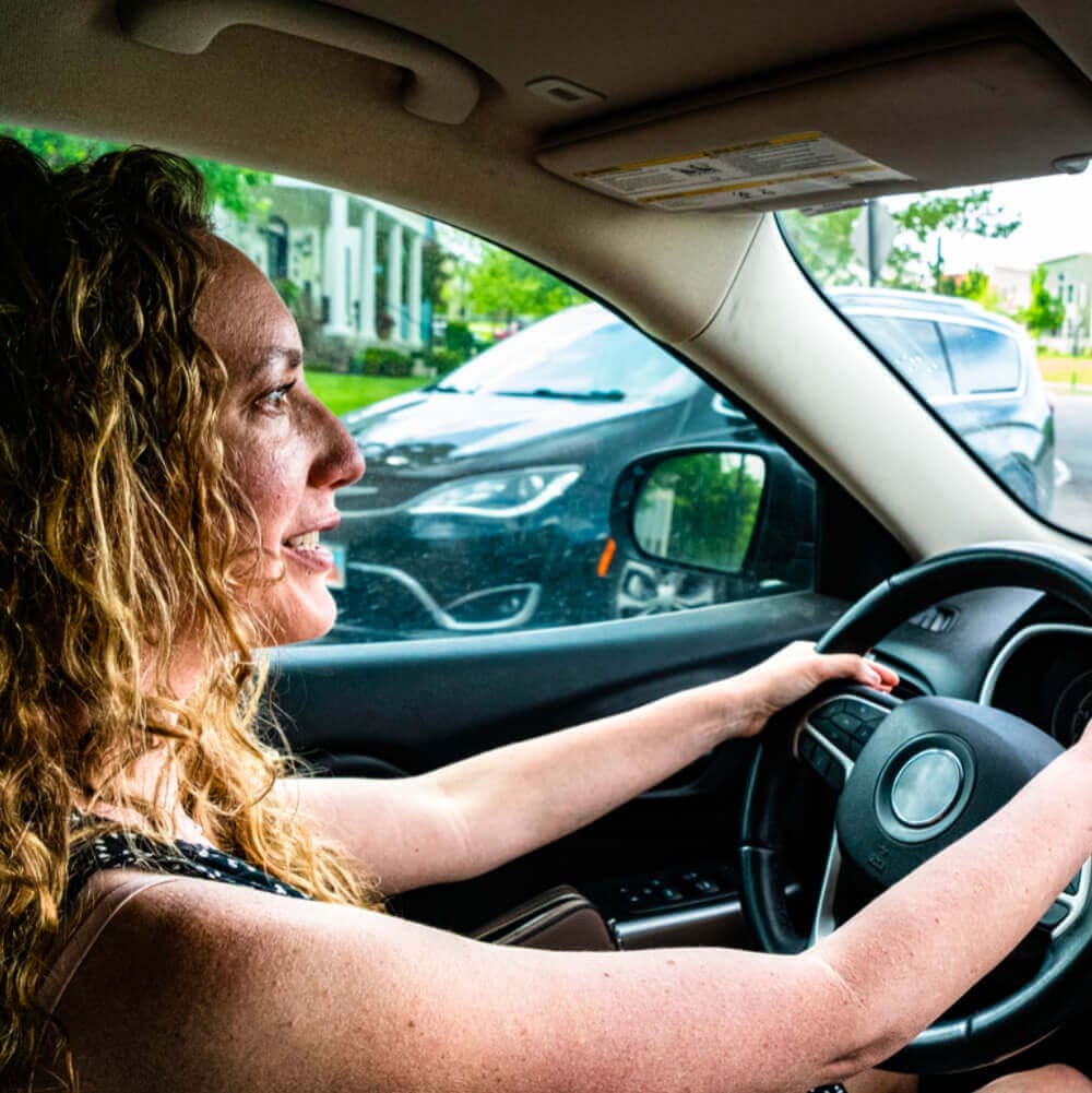 A woman with curly hair is driving a car, smiling as she looks ahead. The well-lit interior reveals a modern dashboard, perhaps hinting at a recent radio replacement. Outside the window, a parked black vehicle and a residential neighborhood with green trees can be seen.
