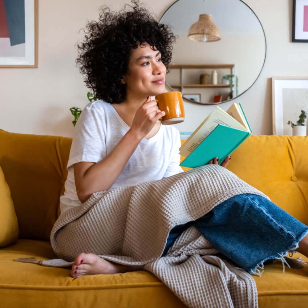 A woman with curly hair sits on a yellow couch, wrapped in a beige blanket. She holds a mug in one hand and a book in the other, smiling and gazing to the side. The room, warmed by her remote start fireplace, features a large mirror and framed pictures on the wall.