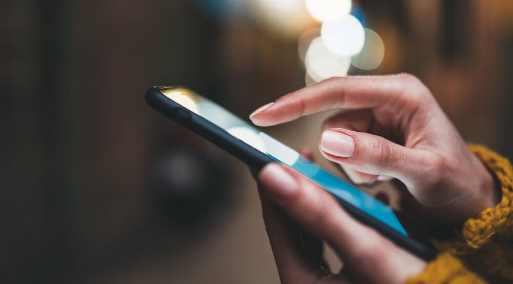 A person in a yellow sweater is using a smartphone, their index finger tapping the screen to activate remote start. The background is an artful blur of circular lights, adding a touch of modernity to the scene.
